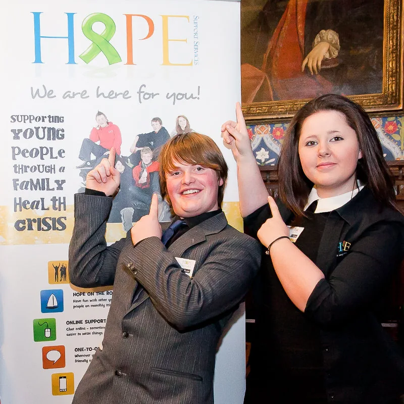 Two young people pointing at a hand-made "We are here for you" support banner.